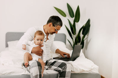 Dad sits on the bed, works on a laptop at home with a baby on his lap, smiles 