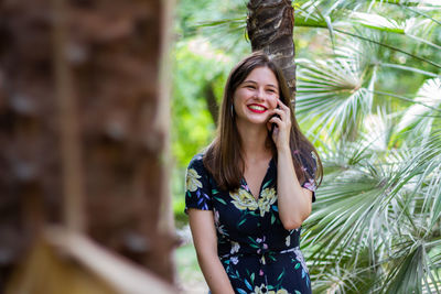 Beautiful young woman using phone while standing against tree trunk