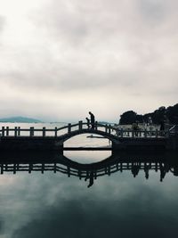 Silhouette man and child walking on arch bridge over lake against sky