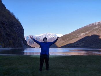 Mature man with arms outstretched standing on grassy field against river