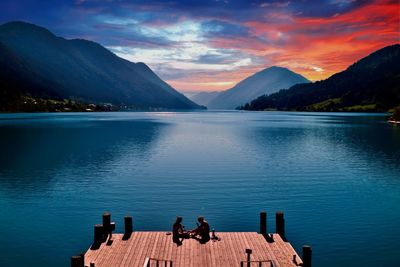 Couple sitting on jetty over lake against sky during sunset