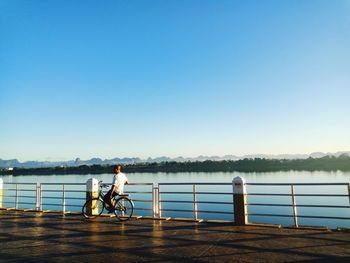 Man with bicycle on railing by lake against clear blue sky