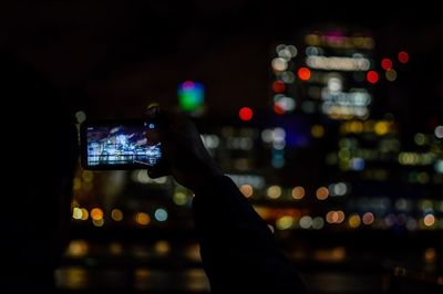 Silhouette hand photographing illuminated city through mobile phone at night