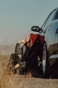 Stylish brunette woman in warm gray coat and knitted burgundy hat is sitting on ground near her car