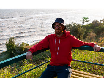 Millennial man in bucket hat red hoody eyeglasses on a bench with sea. authentic tourist lifestyle.