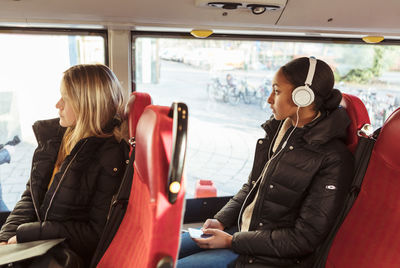 Teenage girl listening music through headphones and female looking through window while traveling in bus