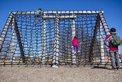 Two small children climb high rope wall outside while father looks on