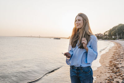 Smiling young woman lwith smartphone standing on the beach