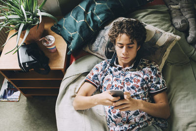 High angle view of male teenager listening music through mobile phone while lying on bed at home