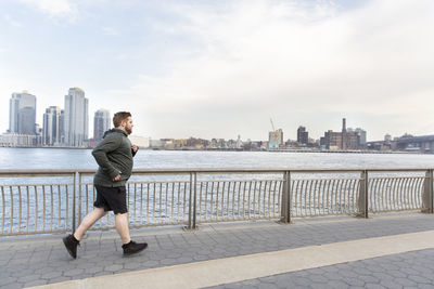 Full length of overweight man jogging on bridge by river in city