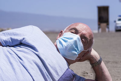 Portrait of senior man wearing flu mask relaxing on beach against sky