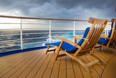Empty wooden lounge chairs on boat deck at sea against cloudy sky