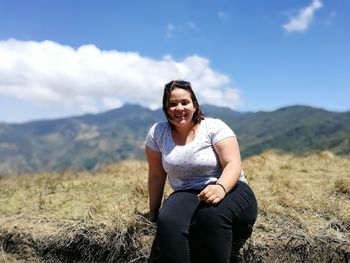 Portrait of smiling young woman sitting on field with mountains in background