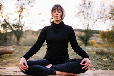 Full length of woman meditating while sitting on bench against trees