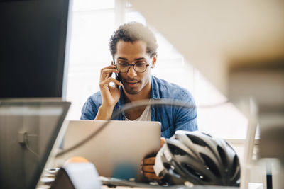 Computer programmer talking on smart phone while using laptop in creative office