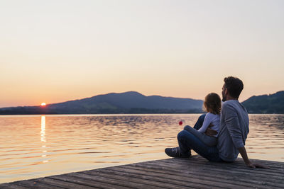 Rear view of man sitting on pier over sea against sky during sunset