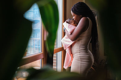 Side view of african american female with long hair standing at window and kissing newborn baby wrapped in soft towel