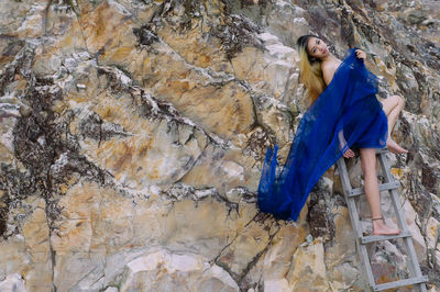 Sensuous woman wrapped in blue fabric leaning on ladder against rock formation