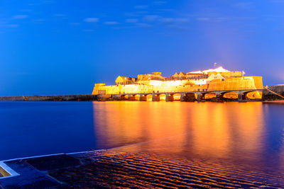 Illuminated buildings by sea against blue sky at night