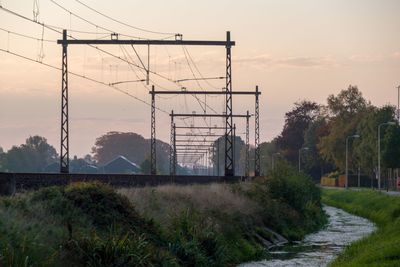 Electricity pylon by trees against sky during sunset