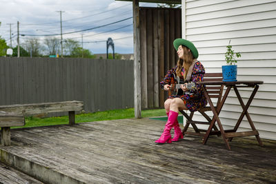 A stylish female singer sits on a deck in a backyard playing guitar