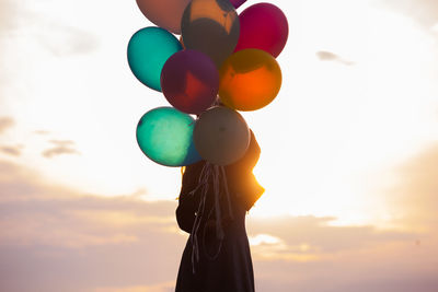 Low angle view of woman holding colorful balloons against sky during sunset