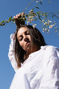 Low angle portrait of young woman holding flowers against clear sky