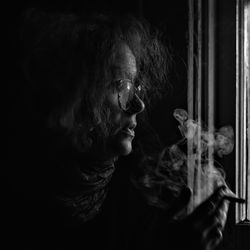 Side view of mature woman smoking while standing by window in darkroom at home