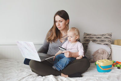 Mother using laptop while sitting with baby girl on bed at home