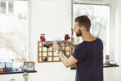 Side view of young man standing against window at home