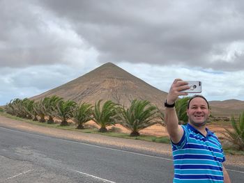 Smiling man taking selfie on mobile phone while standing on road against sky
