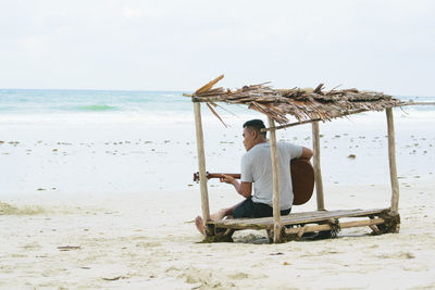 Man playing guitar while sitting under shed at beach