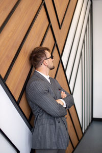 A young man in a suit and sunglasses is standing near the wall with his hands folded