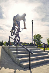 Male rider riding bmx bike on railing against sky at skateboard park during sunny day