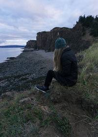 Woman sitting on rock looking at sea shore against sky