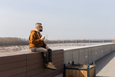 Side view of man sitting on retaining wall against clear sky