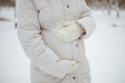 A pregnant woman in a winter jacket and mittens holds her hands on her stomach while waiting 