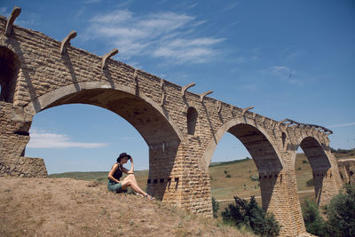 Woman traveler explorer in a black hat sit next to the destroyed old stone bridge in the summer