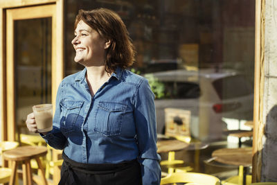 Happy female owner holding coffee cup while standing against cafe