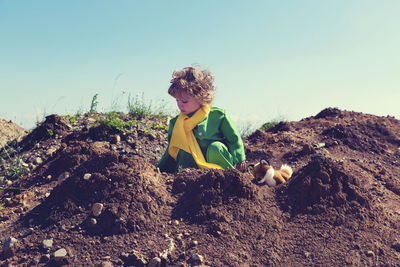 Cute boy crouching on dirt against sky