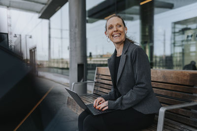 Cheerful businesswoman sitting with laptop on bench while waiting at railroad station