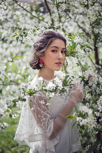 Portrait of a young beautiful woman in white clothes standing next to blooming cherry tree in spring
