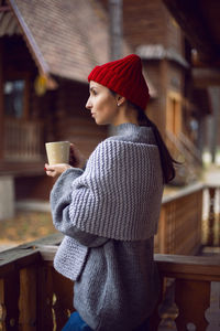 Woman in a red hat and scarf and a mug stands at a wooden house, in the woods in the autumn morning 