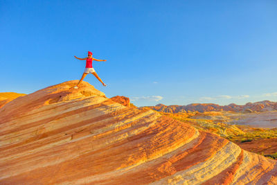 Full length of mature woman jumping on rock formations against blue sky during sunset