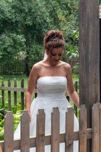 Smiling bride in wedding dress standing by fence against trees