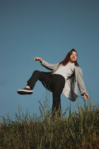 Low angle view of young woman standing on one leg over grass against clear blue sky
