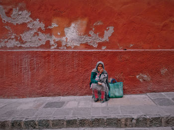 Female beggar sitting against red wall on sidewalk