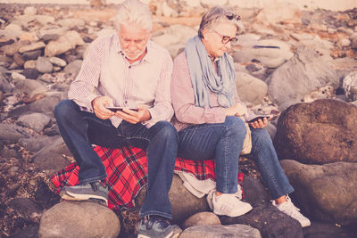 Couple using mobile phones while sitting on rocks at beach