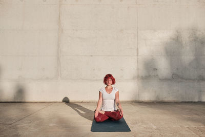 Flexible barefooted woman with red hair and tattoo in white shirt and burgundy pants sitting in padmasana with gyan mudra and meditating with closed eyes against concrete wall on street among urban environment during sunrise