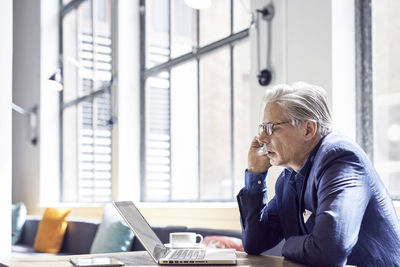 Businessman looking at laptop computer while sitting against windows in office cafeteria